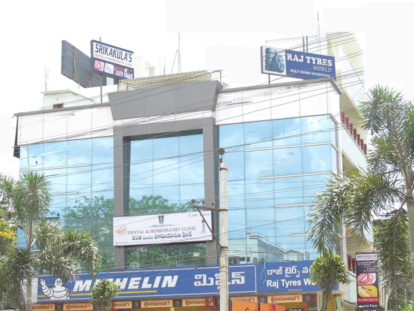 A modern commercial building displaying multiple signs, including "Srikkakulas," "Raj Tyres World," and the best homeopathy clinic in Tirupati. The structure features reflective glass windows and is flanked by tropical trees and various street-level storefronts, including Michelin.