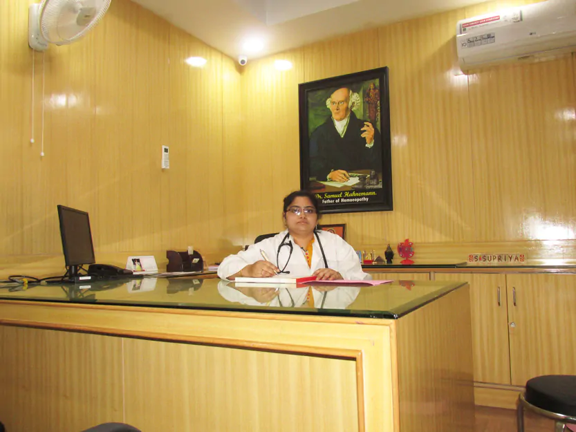 A doctor from Srikakulas Dental Clinic sits at a wooden desk in an office, writing on a clipboard. The room has a wooden interior, with a small computer on the desk and an air conditioner mounted on the wall. A large framed portrait hangs on the wall behind the doctor in this renowned Tirupati clinic.